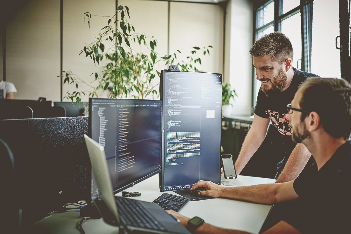 Zwei Männer im Büro besprechen Code auf Monitoren.